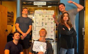 Five people pose in front of a wall chart, one holding a paper, the group appears celebratory. The setting is an indoor office space with the name, ZDIGITAL, above the door.