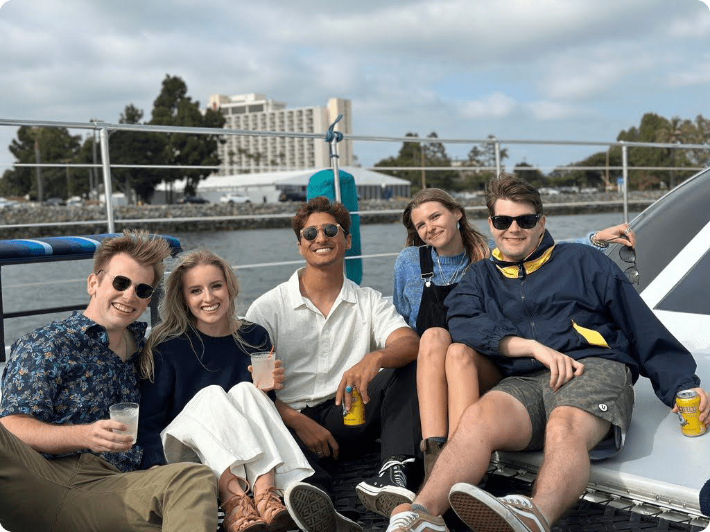 Five people are sitting on a boat, smiling and holding drinks. In the background, water, trees, and buildings are visible under a cloudy sky.