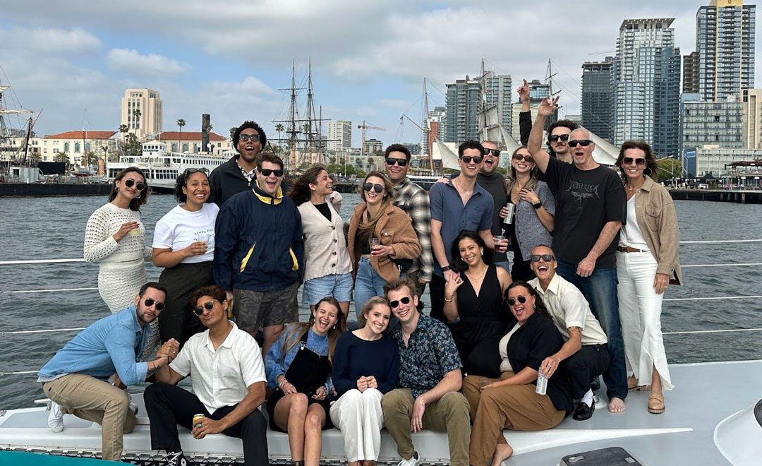 A group of people posing for a photo on a boat with a city skyline in the background. Many are smiling and some are holding drinks. The weather appears to be clear and sunny.