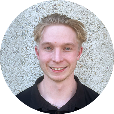 A young man with short, blonde hair smiles in front of a textured, gray wall.