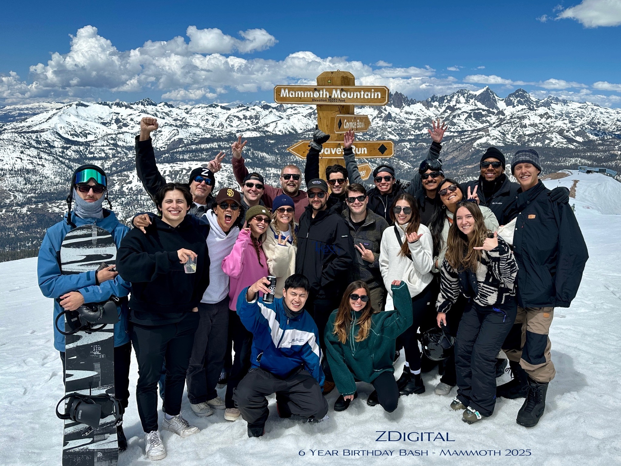 A group of people in ski gear are raising glasses and smiling at an outdoor table near a snowy mountain on a sunny day. Ski lifts and pine trees are visible in the background.