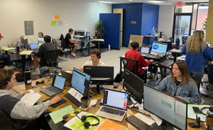 Office with multiple people working at desks, using computers and wearing headsets; papers, monitors, and office supplies are visible throughout the workspace.