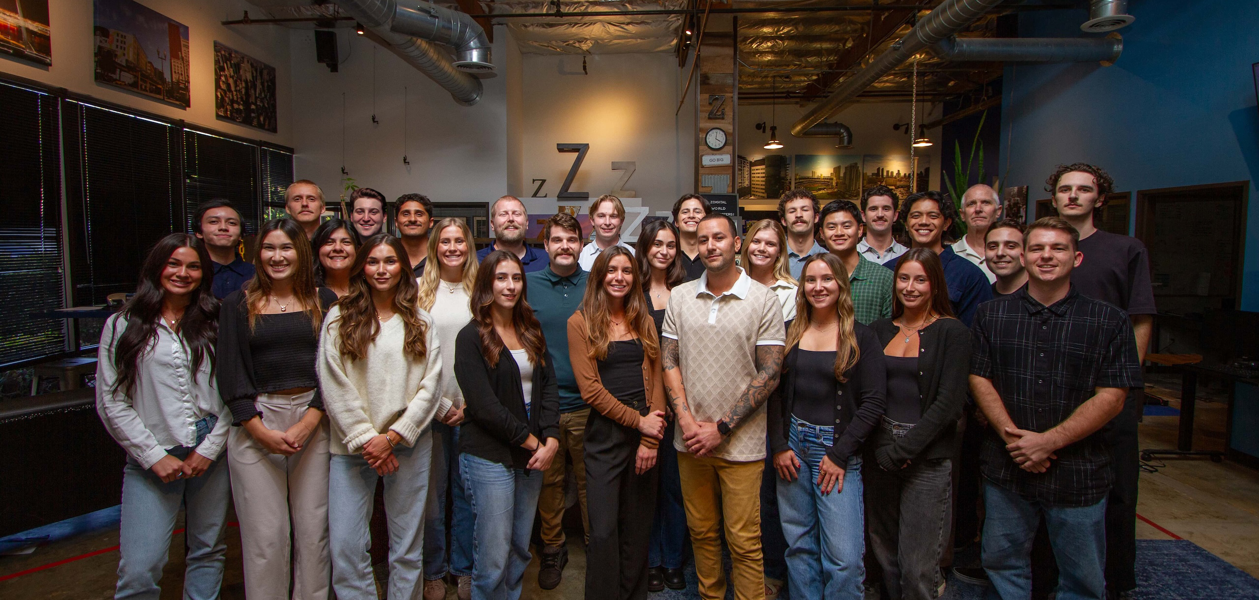 A group of around 30 people posing together indoors in a casual office setting, with art on the walls and visible ceiling pipes above.