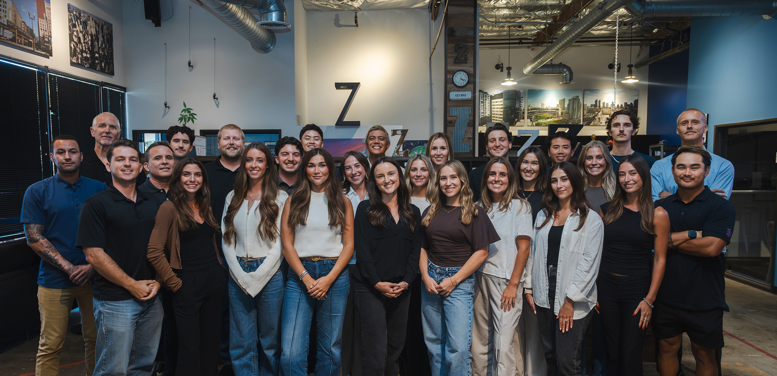 A group of twenty-six adults poses for a photo in an office space with modern industrial decor, including exposed pipes and large wall art.