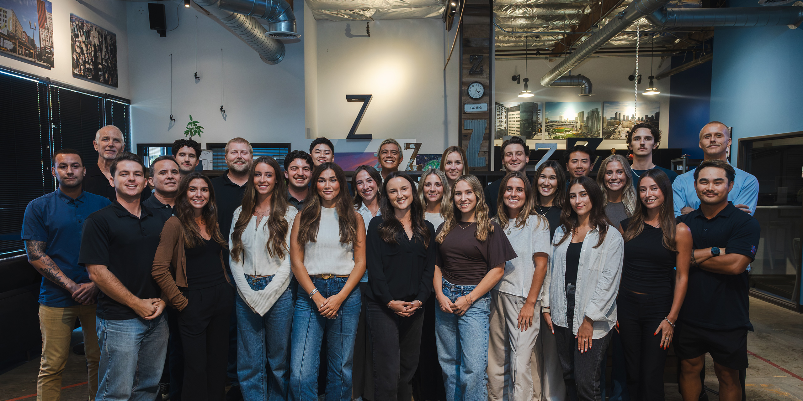A group of men and women stand together indoors, posing for a team photo in a modern office setting with industrial decor.