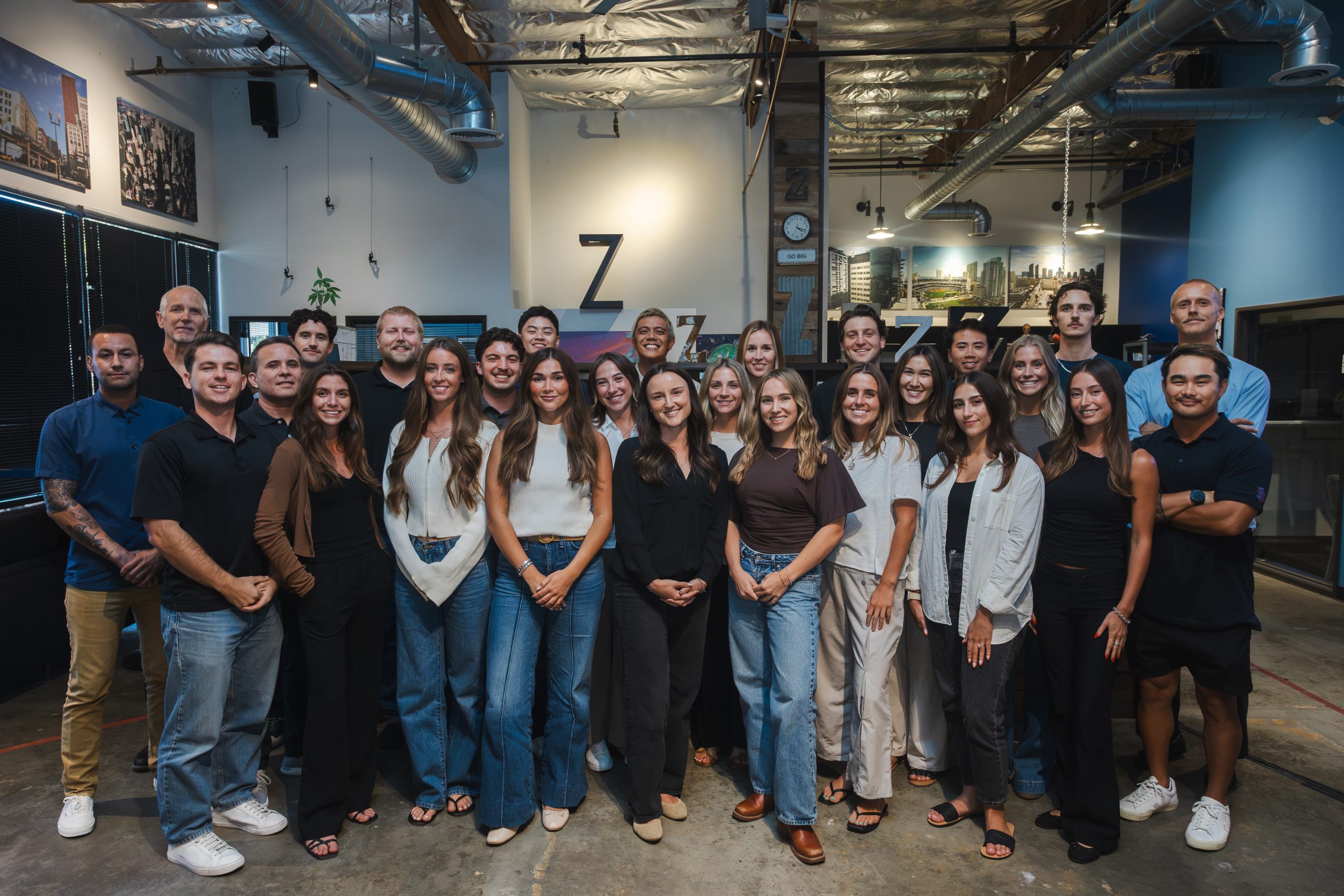 A group of 26 adults pose together inside an industrial-style office space, standing in rows and smiling at the camera.